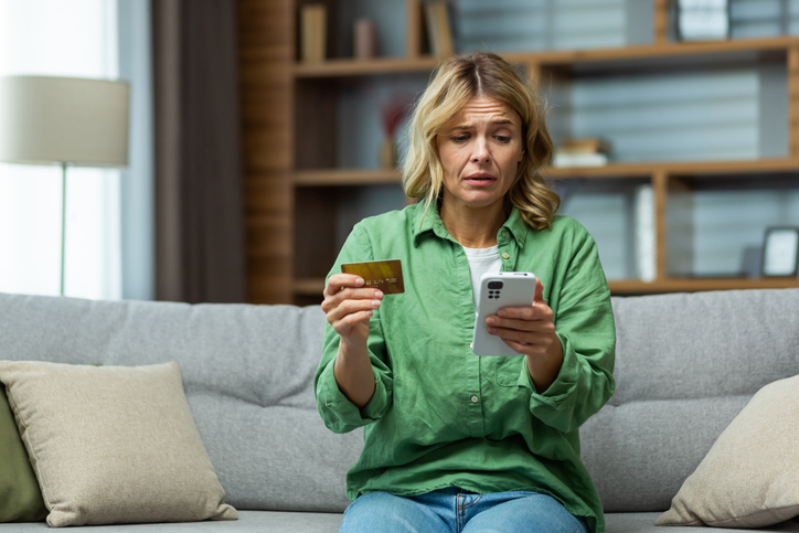 A woman in a green shirt holding a credit card and her phone, hesitating before typing in the card details